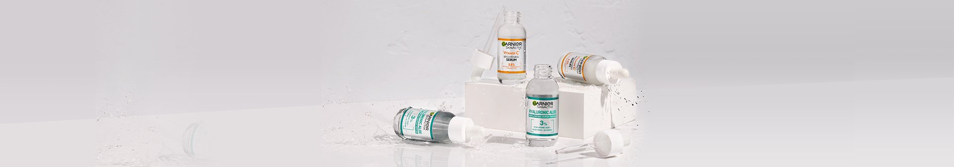 Hero banner with four face serums on a grey background. Serums are two bottles of Vitamin C and two bottles of Hyaluronic Acid.